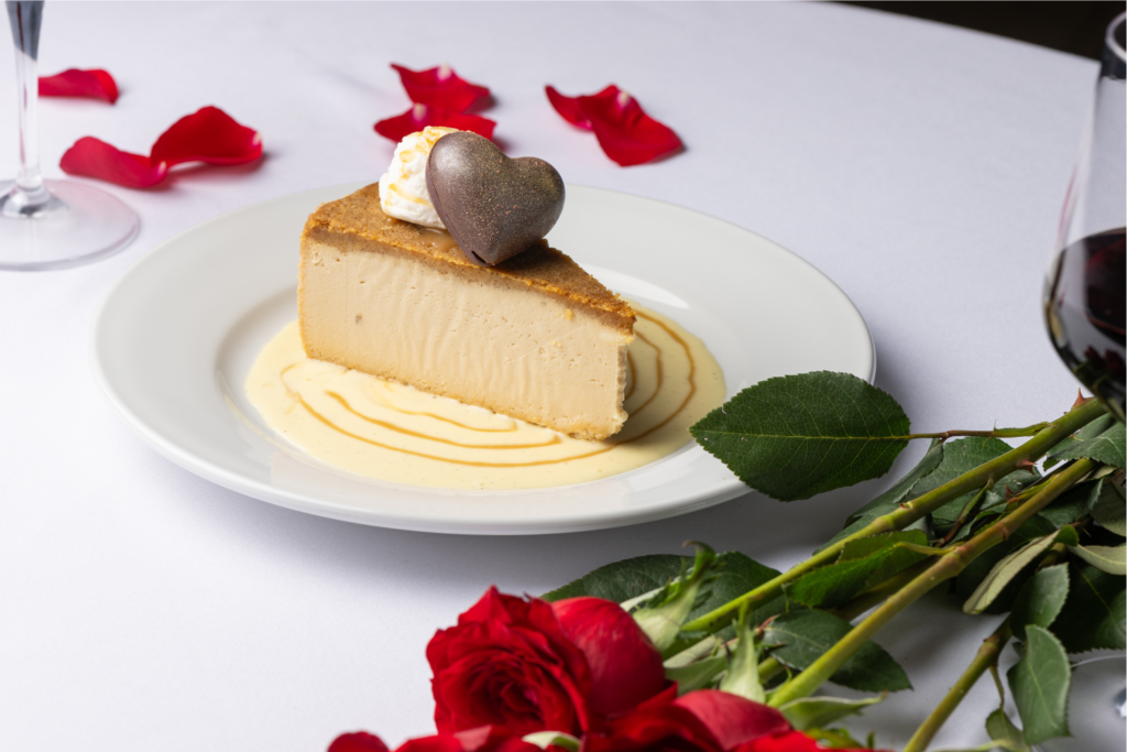 Slice of creamy cheesecake topped with a chocolate heart and whipped cream, served on a white plate with red roses and petals nearby.